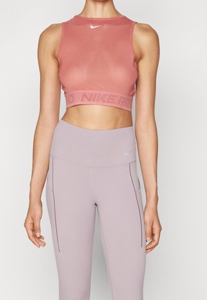 Kobieta ubrana w dopasowany różowy top Nike bez rękawów oraz jasnofioletowe, wysokowalne legginsy Nike, stojąca na jednolitym tle.