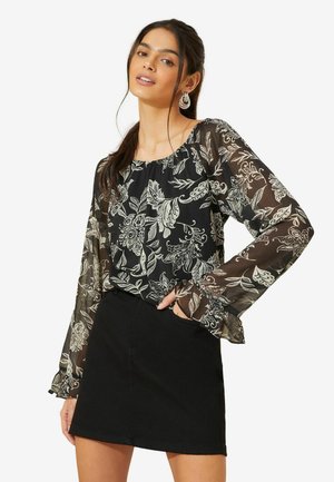 Schwarzblumiges Blusenshirt mit transparenten Ärmeln, versehen mit weißen Blumenmustern und einem runden Halsausschnitt, kombiniert mit einem schwarzen Denim-Rock.