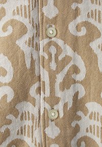Tessuto stampato con un design astratto marrone e bianco e una texture liscia. Un bottone rotondo e leggero è visibile lungo la cucitura.
