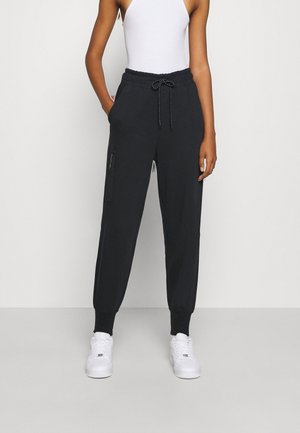 Personne portant un jogging noir avec cordon et poche zippée, un top blanc sans manches et des baskets blanches, debout devant un fond uni.