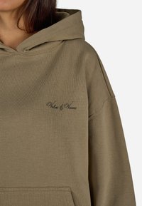 Olivegrüner Hoodie mit einer großen Kängurutasche, verstellbarem Kapuzenzug und gesticktem "Values & Visions"-Logo in Schwarz. Weiche Textur.