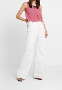 Rosa ärmlös blus med plisserad front och spetsdetaljer, kombinerad med högmidjade vita vida jeans. Svarta sandaler fulländar looken.