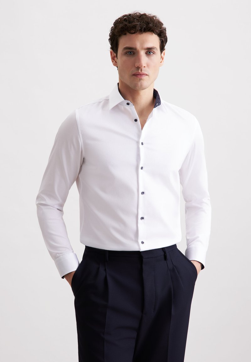 Camicia bianca a maniche lunghe con colletto a fantasia blu e bottoni coordinati, abbinata a pantaloni scuri. Tessuto morbido con un design aderente.