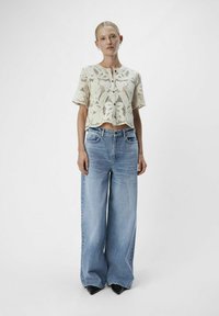 Top corto de encaje en color crema con mangas cortas y cierre de botón, combinado con unos vaqueros de pierna ancha en azul claro y zapatos negros de punta.