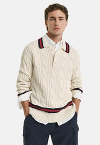 Kabelstrikket cremesweater med polo-krave, der har en mørkeblå og rød stribet kant ved kraven og i kanten. Lange ærmer.