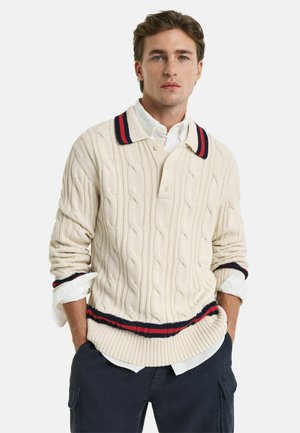 Suéter de punto trenzado crema con cuello tipo polo, con una banda a rayas en azul marino y rojo en el cuello y el dobladillo. Manga larga.