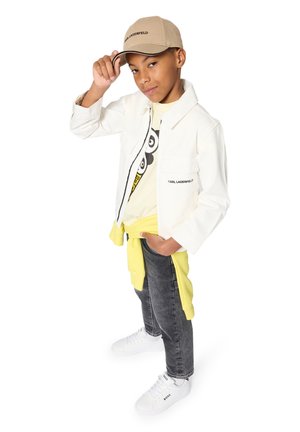 Witte spijkerjas, geel grafisch T-shirt, grijze spijkerbroek, gele trui om de taille gestrikt en witte sneakers met merklogo. Beige pet.