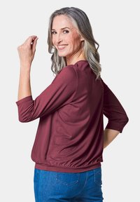Bordeauxkleurige blouse met driekwartmouwen, zachte stof, geplooide zoom en een relaxte pasvorm, gecombineerd met blauwe jeans.