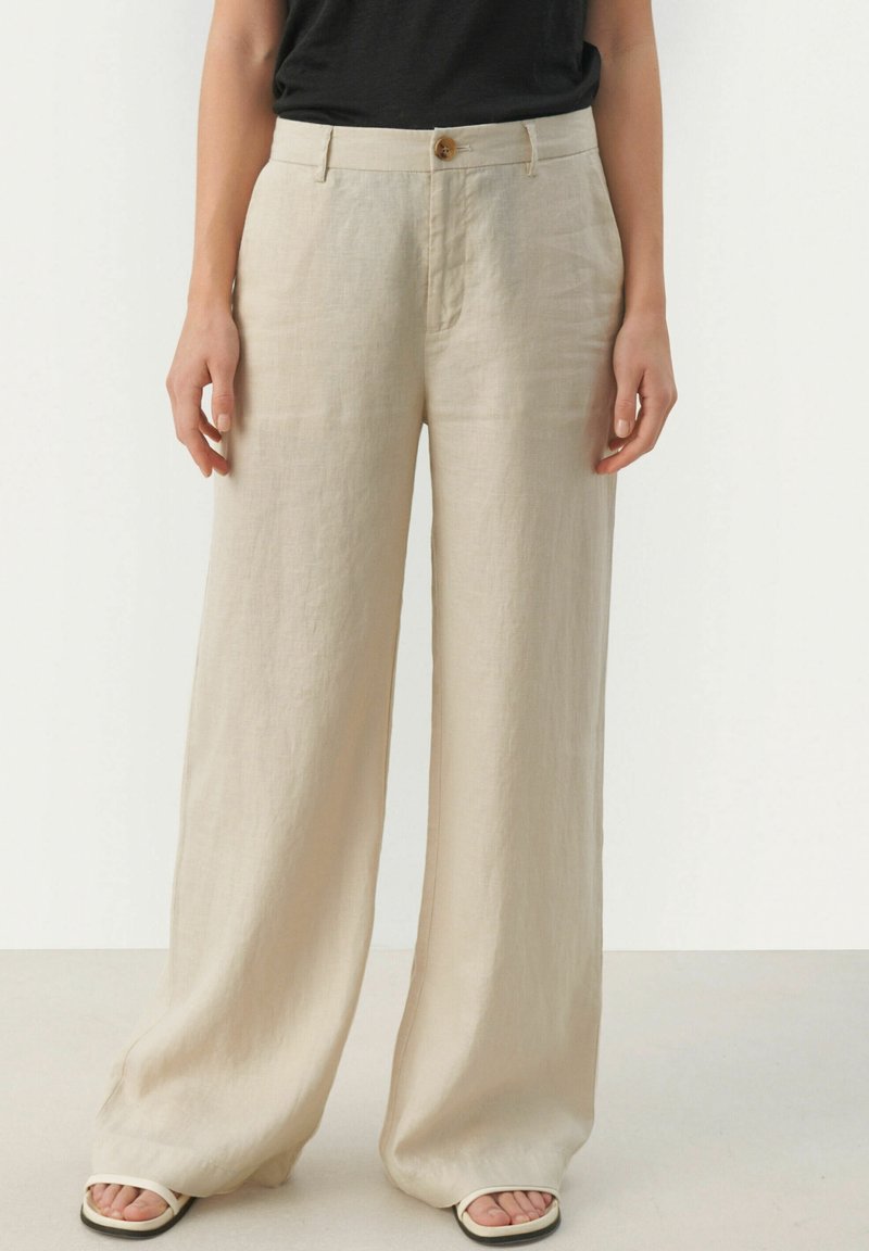 Pantalon en lin beige taille haute à jambes larges avec bouton et fermeture éclair à l'avant, porté avec un haut noir et des sandales blanches à bouts ouverts.