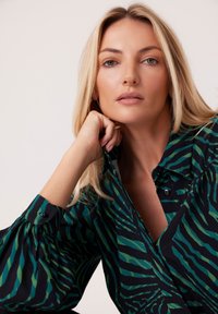 Czarna koszula w zielone paski w zebrę, z guzikiem z przodu, długimi rękawami z plisowanymi mankietami i złożonym kołnierzykiem. Gładka faktura materiału.