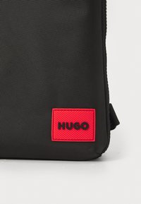 Schwarze, strukturierte Stofftasche mit einem auffälligen roten Gummilogo-Patch mit der Aufschrift "HUGO". Hat ein elegantes, modernes Design.