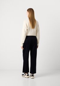 Vrouw die van zich af kijkt in een crèmekleurige trui, zwarte wijde broek en zwart-witte sneakers tegen een effen witte achtergrond.
