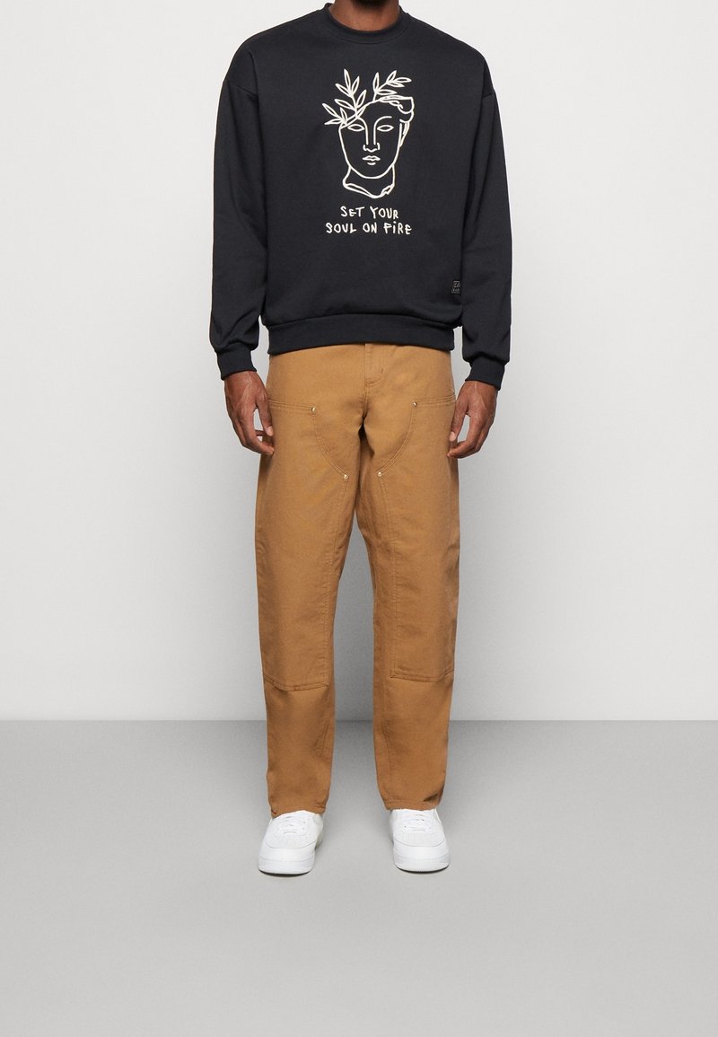 Sudadera negra con diseño de arte lineal en blanco y texto, combinada con pantalones beige de corte holgado que cuentan con bolsillos y costuras sutiles. Zapatillas blancas.