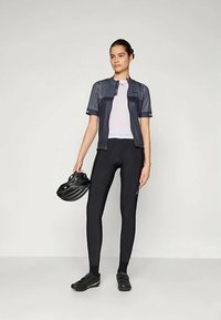 Μαύρα κολάν ποδηλασίας συνδυασμένα με ένα σκούρο γκρι κοντομάνικο μπλουζάκι και μια λευκή μπλούζα εσωτερικά. Ένα μαύρο κράνος κρατείται με το αριστερό χέρι.