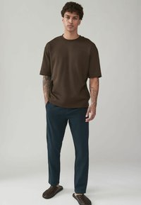 Brown short-sleeve t-shirt with a round neckline, paired with dark blue tailored pants and dark woven slide sandals.