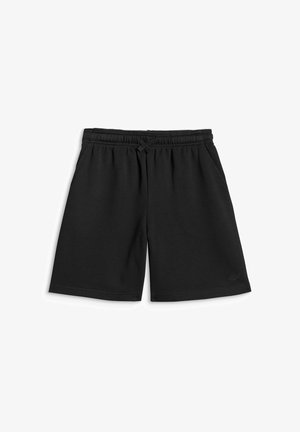 Garçon portant un short noir et un t-shirt noir avec un texte orange, tenant une veste noire, debout devant un fond gris uni.