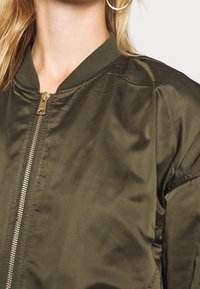 Olivově zelená bomber bunda s lesklou texturou, žebrovaným límcem a zlatým zipem. Disponuje klasickým, svobodným designem a jednolitou barvou.