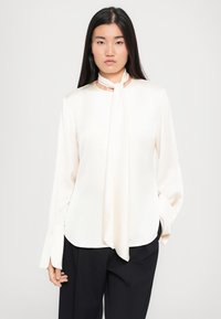 Bluză crem cu o textură moale, guler rotund, mâneci lungi cu o fante și un accent de eșarfă legată la gât.