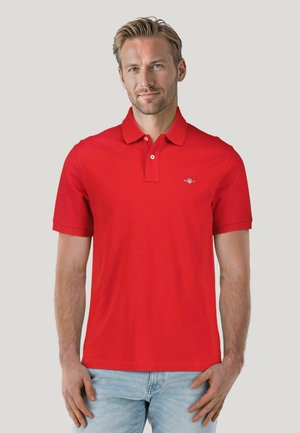 Red polo shirt with a ribbed collar, two-button placket, and small logo on the chest. Short sleeves and straight hem. Cotton material.