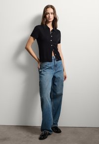 Eine Frau steht an einer weißen Wand und trägt ein schwarzes kurzärmliges Button-Up-Oberteil, lockere blaue Jeans und schwarze Loafers, mit einer Hand in der Tasche.
