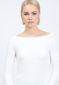 Witte, langärmelige top met een off-the-shoulder halslijn. Zachte, lichtgewicht gebreide stof met minimale naden. Eenvoudige textuur, geen patronen of accenten.