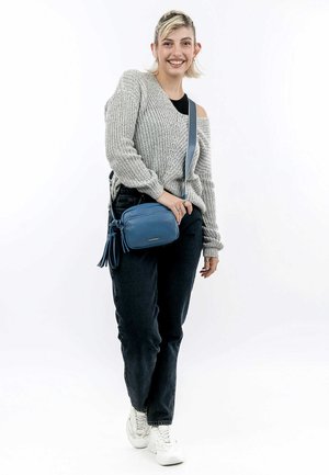 Blue leather crossbody bag with a textured finish, adjustable strap, and decorative tassels. Model wears a gray knitted sweater and dark jeans.
