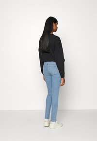 Sweatshirt preto cropped, jeans skinny azul claro, ténis brancos. Modelo visto por trás contra um fundo branco liso.