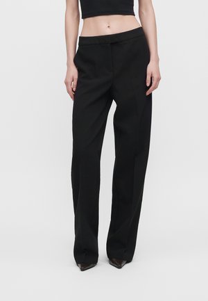 Modèle portant un pantalon noir ample à jambes larges et à la ceinture asymétrique, un crop top noir et des chaussures pointues, sur un fond clair uni.