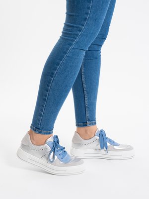 Legs wearing blue skinny jeans and white sneakers with blue laces, standing on a white background.