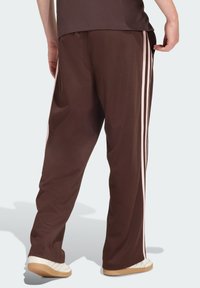 Brązowe spodnie sportowe z różowymi paskami po bokach, elastyczną talią i gładką fakturą. Stylizowane z białymi butami.