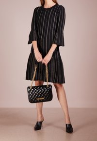 Femme en robe noire à rayures verticales dorées, tenant un sac à main noir matelassé avec des chaînes dorées, et portant des mules noires pointues.