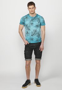 Zila ziedu raksta T-kreklu ar apaļu kakla izgriezumu, kombinēts ar melniem džinsa šortiem. Šortiem ir sarullēti mali un priekšējās kabatas; modele valkā tumšus sporta apavus.