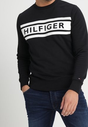 Mężczyzna noszący czarną koszulkę z długim rękawem z białym prostokątnym logo z napisem "HILFIGER" na piersi i ciemnoniebieskie dżinsy, z jedną ręką w kieszeni.