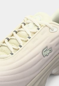 Gros plan sur une basket beige avec mesh respirant, languette rembourrée, lacets croisés et logo crocodile sur le côté.