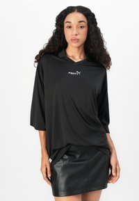 Mladá žena s kučeravými vlasmi, oblečená v voľnej čiernej košeli značky Fenty a čiernej koženej mini sukni na jednoduchom bielom pozadí.