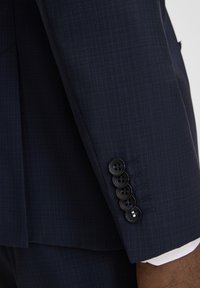 Nahaufnahme eines marineblauen Blazers mit strukturierter Stoffoberfläche, figurbetontem Design und fünf schwarzen Knöpfen am Ärmel mit feinen Steppnähten.