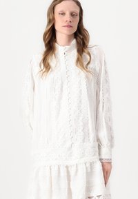 Hvit langermet bluse med teksturerte mønstre, med høy krage, knappefront og intrikate blonder. Mykt, lett stoff.