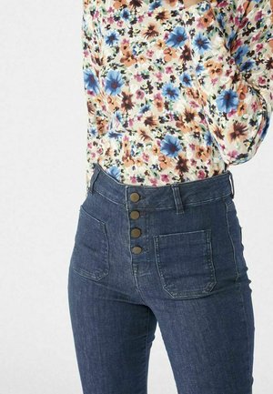 Frau trägt blaue Jeans mit hoher Taille, vorne mit Taschen und Knöpfen, kombiniert mit einer langärmligen, floralen Bluse in Pastellfarben.