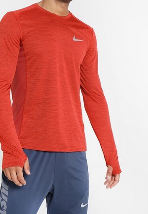 Mann trägt ein rotes langärmliges Nike Sportshirt und blaue Nike Shorts mit sichtbaren Markenlogos auf Brust und Bein.