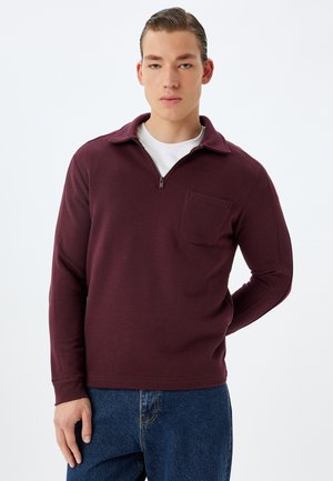 Pullover bordeaux a maniche lunghe con zip metà, dotato di collo e tasca sul petto. Tessuto morbido con vestibilità regolare, abbinato a jeans blu.