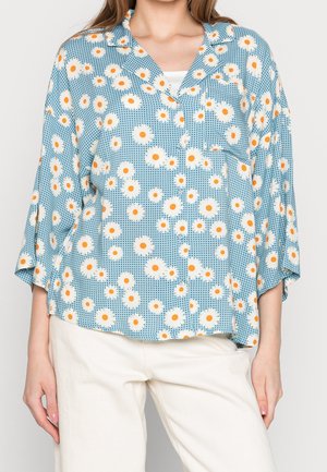 Blouse légère à manches courtes en tissu bleu avec un motif floral blanc et orange, présentant un devant boutonné et une poche à rabat.