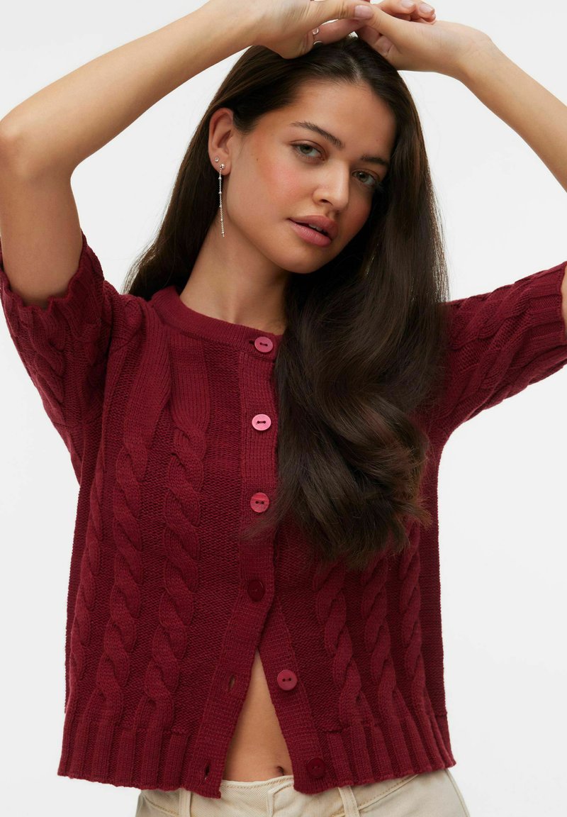 Femme aux longs cheveux foncés portant un cardigan bordeaux en maille torsadée boutonné et un pantalon beige, levant les bras au-dessus de sa tête.