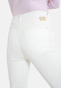 Jean en denim blanc avec un design ajusté, comportant des poches arrière, des détails de couture subtils et une étiquette en cuir tan près de la taille.