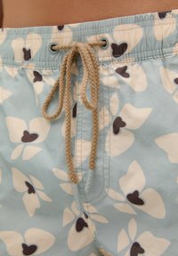 Shorts de bain bleu clair avec des motifs floraux en forme de cœur blancs et marron foncé. Présente une taille à cordon de serrage et une texture tissée.