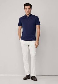 Tmavě modrá polo košile s krátkými rukávy, plochým límcem a jednoduchým designem, kombinovaná s bílými kalhotami a tmavě hnědými botami.