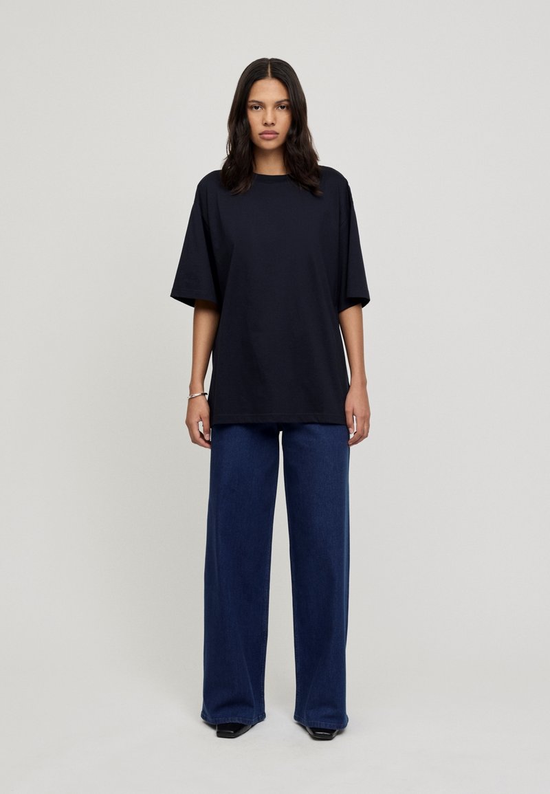 T-shirt oversize bleu marine à manches courtes, associé à un pantalon en denim foncé à jambes larges et des chaussures à talons noires dans un cadre minimaliste.