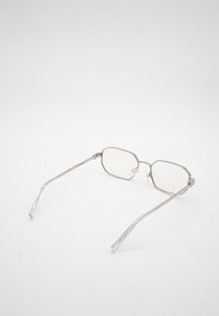 Ορθογώνια γυαλιά με ασημί μεταλλικό σκελετό, διάφανα φακούς και λεπτά μπράτσα, σε λευκή επιφάνεια.