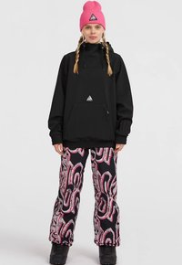 Young woman with two braids wearing a pink beanie, black hooded jacket, and patterned black and pink snow pants with black boots.