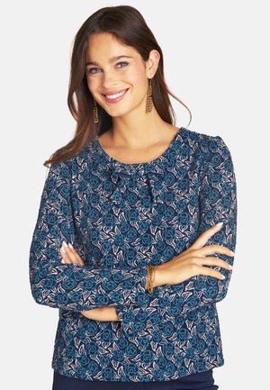 Lächelnde Frau mit langen braunen Haaren trägt ein blaues Langarmshirt mit Blumenmuster, goldene Ohrringe und Armbänder, die Arme verschränkt.