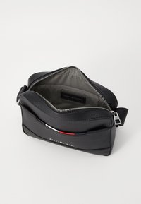 Sac en cuir noir à porter en bandoulière, avec une fermeture éclair, un intérieur gris et un accent à l'avant comportant des rayures rouges, blanches et bleues. Quincaillerie en acier inoxydable visible.